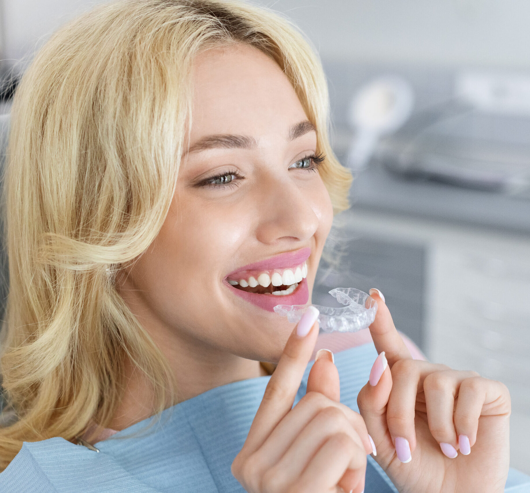 Invisalign orthodontics, modern dentistry concept. Happy young attractive blonde woman holding invisible braces or trainer while sitting at dental chair, closeup portrait, copy space
