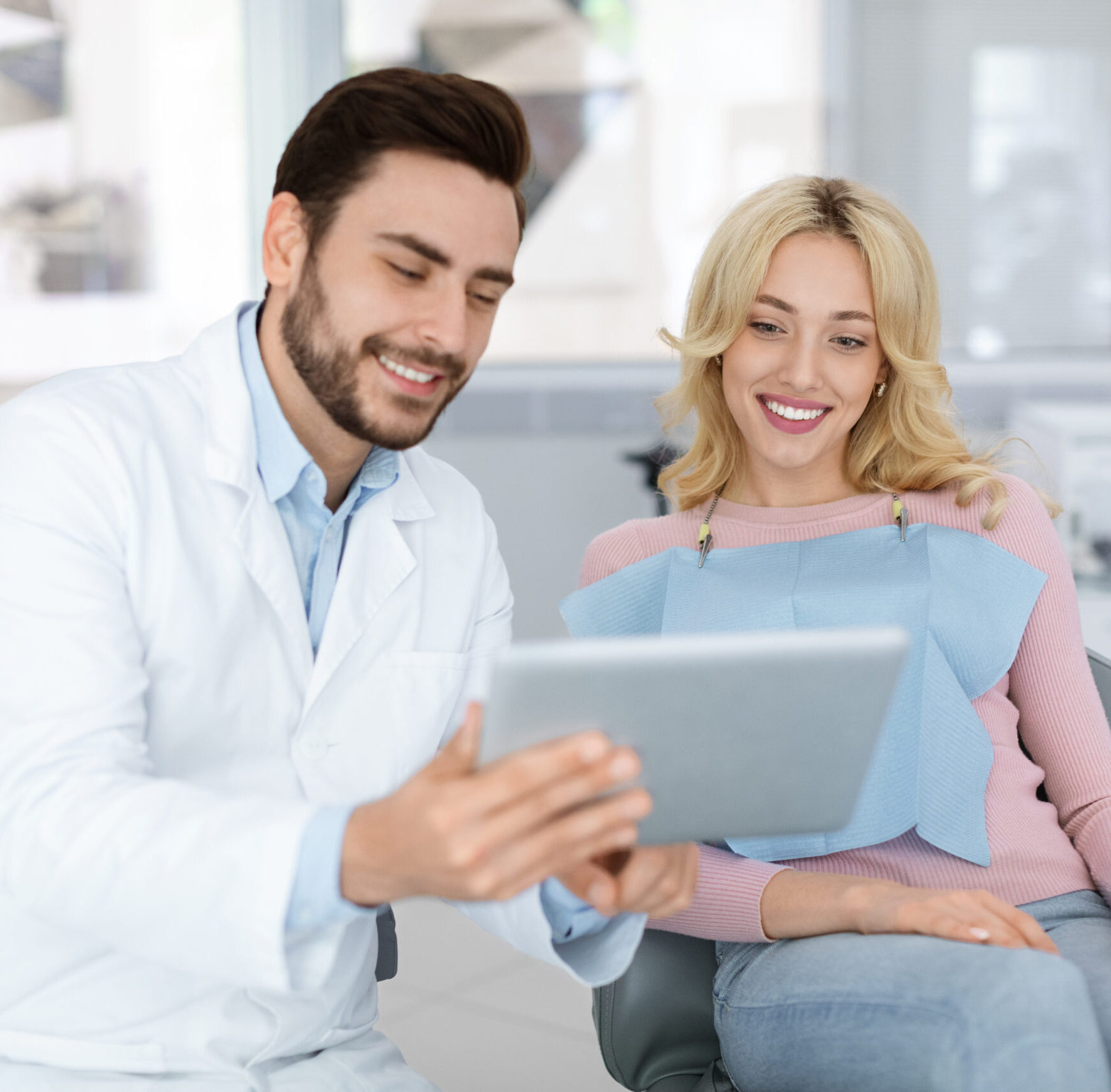Cheerful young bearded man dentist discussing treatment method with beautiful blonde lady patient sitting at dental chair, looking at digital tablet. Modern technologies and healthcare concept